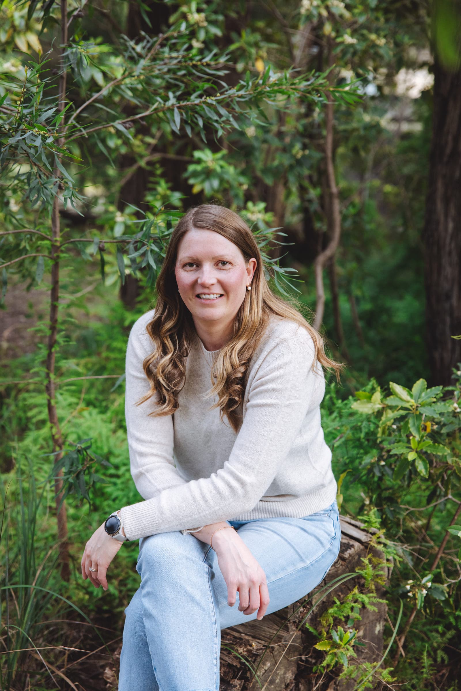 Alisha Perry sitting on a tree stump among the trees near her home, laughing, in a cream knit and jeans