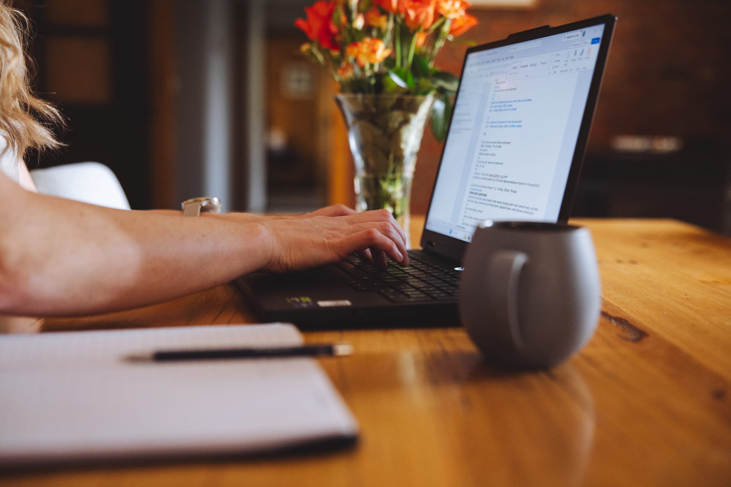 Alisha's hands on her laptop at the kitchen table, with coral roses, a notebook and a mug